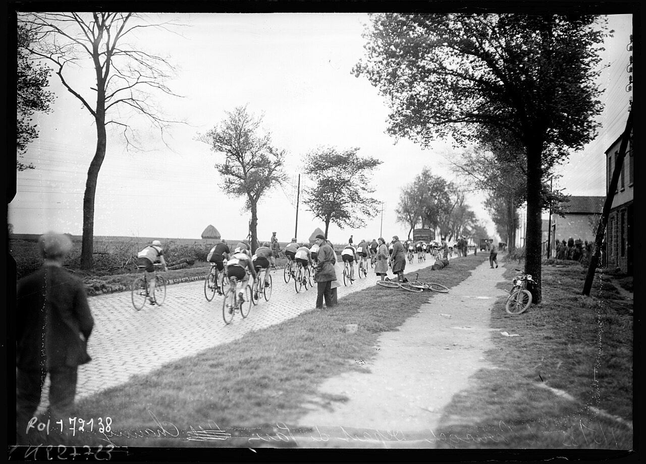 Le peloton s'éloigne sur une route pavée bordée d'arbres au départ de l'édition 1933